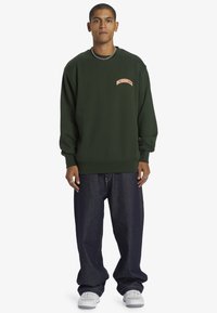 Sudadera verde oscura con cuello redondo y logo en texto naranja. Combinada con vaqueros azules de corte holgado y zapatillas blancas, con un accesorio de cadena.