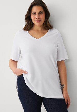 Kvinde med mørkt hår iført en hvid V-hals kortærmet top og mørkeblå jeans, stående med den ene hånd i lommen mod en hvid væg.