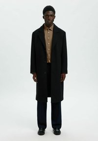 Μαύρο μάλλινο παλτό με γιακά με σχισμή, κλείσιμο με κουμπιά, φορεμένο πάνω από καφέ πουκάμισο με κουμπιά, συνδυασμένο με μπλε-σκούρο παντελόνι και μαύρα παπούτσια.