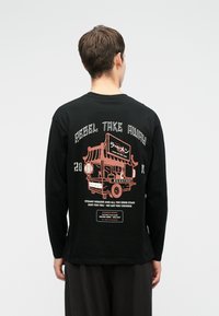 Bărbat îmbrăcat cu o cămașă neagră cu mâneci lungi, având un grafic cu un cărucior de mâncare roz și textul "Rebel Take Away" plus alte inscripții mai mici pe spate.