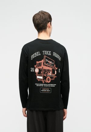 Mężczyzna w czarnej koszuli z długim rękawem, na której znajduje się różowa grafika wózka z jedzeniem oraz napis "Rebel Take Away" z dodatkowymi małymi nadrukami z tyłu.