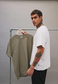 Jeune homme avec des tatouages tenant un t-shirt vert olive sur un cintre devant un portant à vêtements sur fond gris uni.