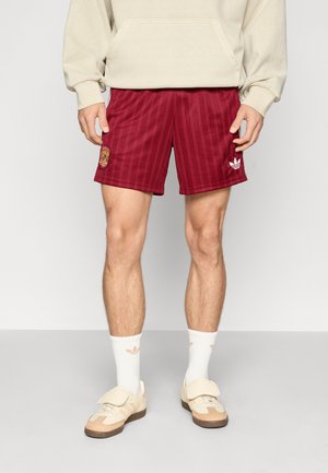 Shorts de sport bordeaux avec des rayures verticales, taille élastique, écusson brodé et logo. Associés à des chaussettes blanches et des baskets beiges.