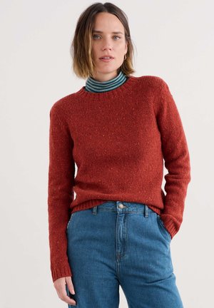 Frau mit schulterlangem braunem Haar, die einen rostfarbenen Strickpullover über einem türkis- und schwarzgestreiften Rollkragenoberteil und blauen Jeans trägt.
