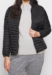 Giacca imbottita nera con zip frontale, colletto alla coreana e polsini elasticizzati. Abbinata a un maglione grigio chiaro lavorato a maglia e pantaloni a quadri.