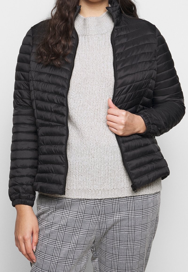 Giacca imbottita nera con zip frontale, colletto alla coreana e polsini elasticizzati. Abbinata a un maglione grigio chiaro lavorato a maglia e pantaloni a quadri.