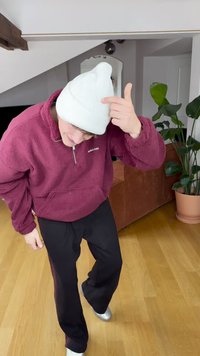 Bordó polár pulóver fél cipzárral, fekete nadrág és fehér kötött sapka. Fa padlón áll egy növény és egy barna kanapé közelében.