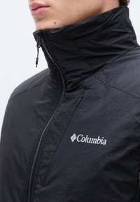 Közeli kép egy személyről, aki fekete Columbia dzsekit visel, amely fel van húzva a nyakáig, a mellkason jól látható logóval.