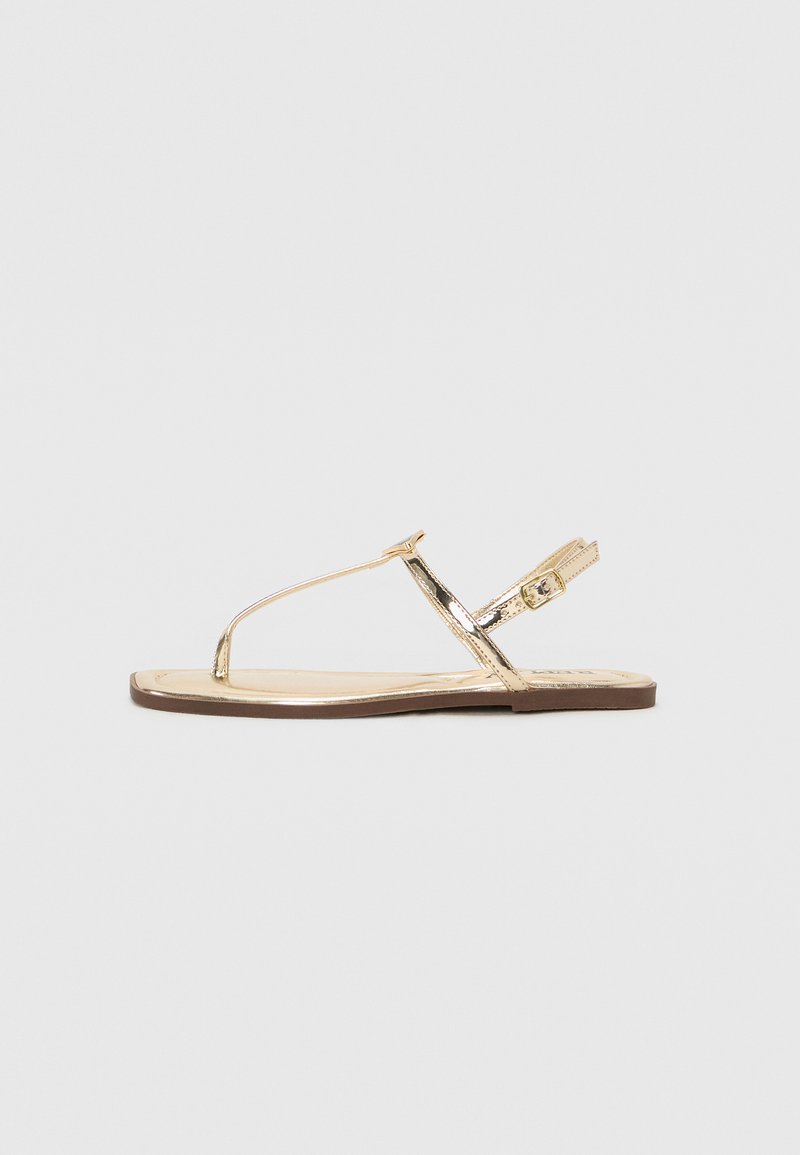 Gold metallic flat sandal with thin straps, adjustable buckle, and a smooth sole. Features a simple design and minimalist style.