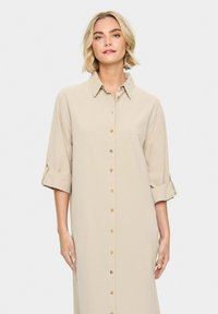 Rochie camasa din material bej, cu guler, maneci lungi suflecate si butoane decorative pe fata. Textura moale.