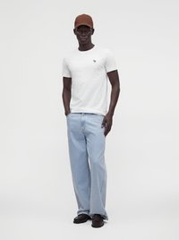 Homme debout portant un t-shirt blanc, un jean large bleu clair, des chaussures noires et une casquette marron sur un fond uni clair.