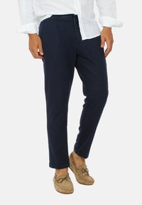 Pantaloni blu navy realizzati in tessuto leggero con un design a gamba affusolata, dotati di pieghe frontali e una cintura elastica. Abbinati a scarpe beige.