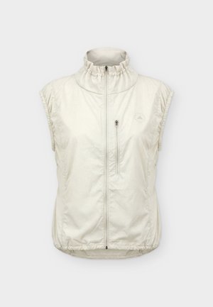 Jeune personne debout, portant une veste beige sans manches, des leggings gris, des chaussettes blanches et des chaussures de sport blanches, sur un fond clair uni.