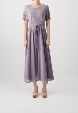 Žena v levandulových krajkových šatech s výstřihem do V, krátkými rukávy, páskem vázaným v pase a splývavou midi sukní z šifonu, doplněná béžovými podpatky.