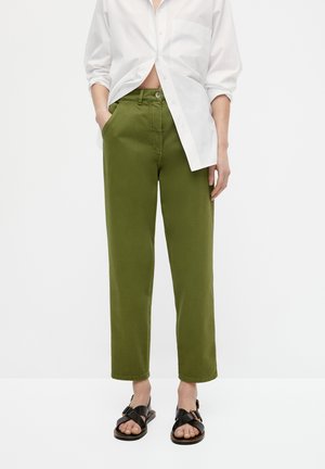 Femme portant un pantalon taille haute vert olive, une chemise blanche boutonnée partiellement sortie, et des sandales noires ouvertes à brides avec boucle.