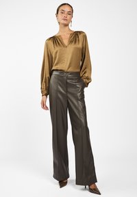Olijfgroene, glanzende blouse met een V-hals en pofmouwen, gecombineerd met donkerbruine, wijde leren broek en puntige hakken.