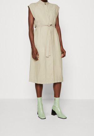 Robe midi beige sans manches avec ceinture nouée à l'avant, associée à des bottines vert clair brillantes à talon bloc, portée par une personne debout.