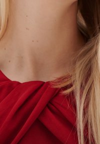 Czerwona bluzka z węzłem przy dekolcie, gładka tkanina i zwiewny fason, oprawiona jasnoblond włosami.