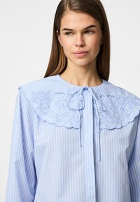 Ανοιχτό μπλε μπλούζα με λευκές ρίγες, διαθέτει κεντημένο κολάρο με γαζιά και φιόγκο στον λαιμό. Μακριά μανίκια, απαλό ύφασμα.