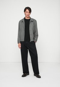 Gray textured jacket with a zip closure, paired with a black T-shirt and loose black trousers. Model stands on a plain background.