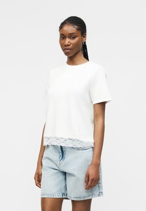 Kobieta z warkoczami, ubrana w biały top z krótkim rękawem i koronkowym wykończeniem oraz jasnoniebieskie jeansowe szorty, stojąca na jednolitym tle.