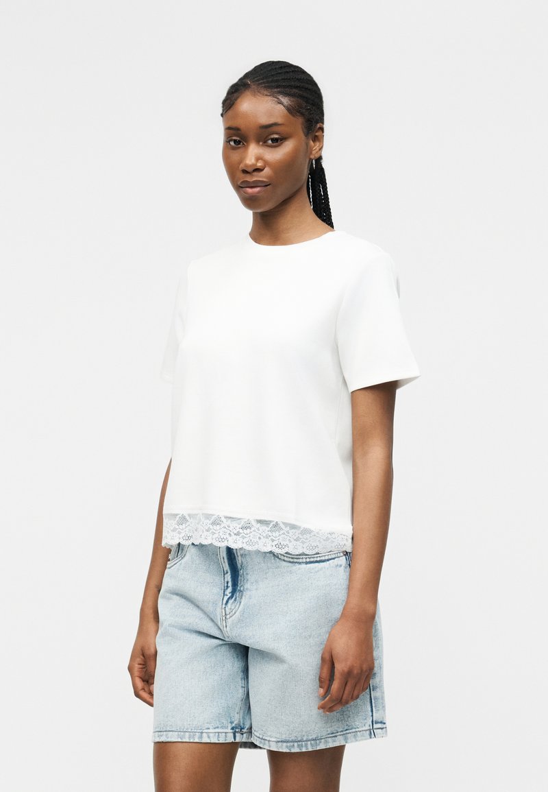 Γυναίκα με πλεξούδες στα μαλλιά, που φοράει λευκό κοντομάνικο τοπ με δαντελένιο τελείωμα και ανοιχτό μπλε τζιν σορτς, στέκεται μπροστά σε απλό φόντο.