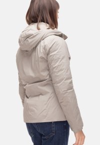 Giacca beige imbottita con cappuccio, caratterizzata da una texture liscia e cuciture lungo la schiena. Abbinata a jeans blu scuro.