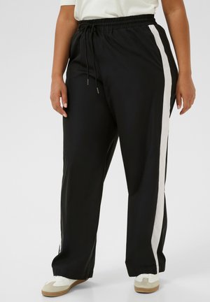 Pantalon noir avec une taille élastique à cordon, bande blanche sur les côtés, forme fuselée, coupe décontractée et tissu doux. Associé à des chaussures blanches et beiges.