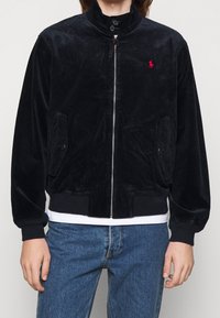 Black corduroy jacket with high collar, zipper closure, two front pockets, ribbed cuffs, and hem. Features a small red logo on the chest.