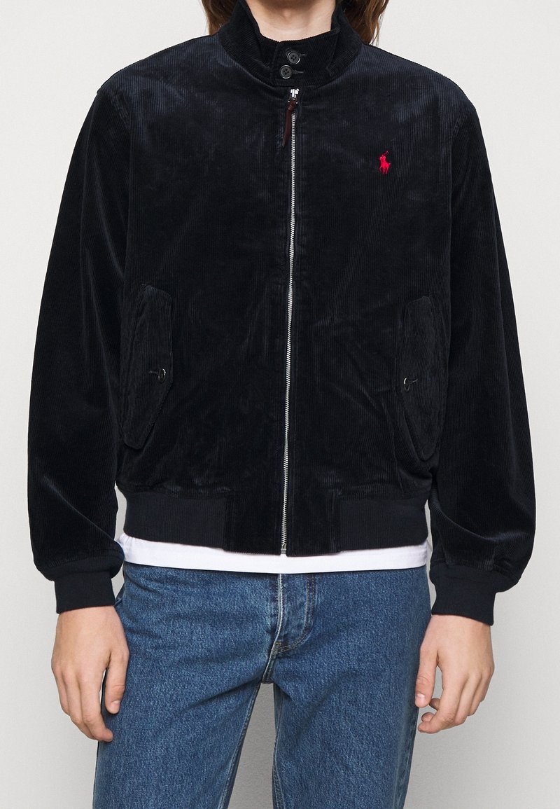 Black corduroy jacket with high collar, zipper closure, two front pockets, ribbed cuffs, and hem. Features a small red logo on the chest.