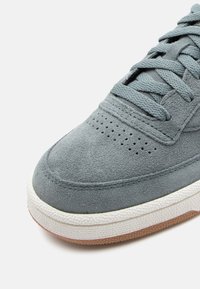 Gaišpelēka zamšota ikdienas apavi ar noapaļotu degunu, perforētu augšdaļu un teksturētu baltu starpsumma, akcentēti ar gumijas zoli.