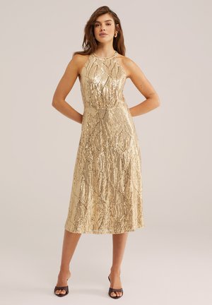 Naine kannab kuldseid litrilisi varrukateta miiidikleit, millel on halteri kaelus, seisab käed selja taga ja jalas on mustad avatud varbaga kontsakingad.