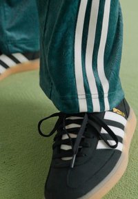 Black athletic sneakers with white stripes and textured detailing. Green pants with three white side stripes. Focus on footwear and pants.