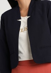 Blazer court bleu marine avec col ouvert minimaliste, deux poches avant et épaules structurées, conçu pour une coupe ajustée.