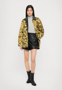 Giacca leopardata giallo e nera con colletto verde, dolcevita grigio, pantaloni corti in pelle neri e stivaletti neri alla caviglia.
