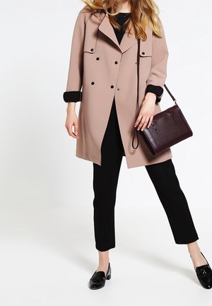 Kobieta ubrana w beżowy dwurzędowy płaszcz, czarne spodnie, czarne buty, trzymająca ciemnobrązową torebkę na ramię.