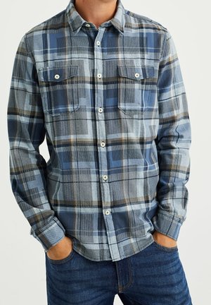 Homme portant une chemise à carreaux bleue et grise à boutons, manches longues et deux poches poitrine, les mains dans les poches d'un jean bleu.