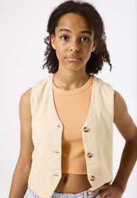 Chaleco de lino beige con botones redondos, colocado sobre una camiseta de tirantes naranja acanalada. Textura suave y ajuste casual, complementado con pantalones cortos de mezclilla claros.