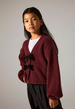Bordeauxrode cardigan gemaakt van zachte stof, met oversized mouwen en twee zwarte fluwelen strikjes aan de voorkant. Witte blouse eronder.