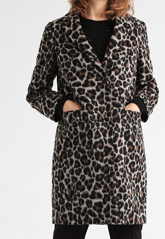 Mujer vistiendo un abrigo de leopardo de largo hasta la rodilla con solapas y manos en los bolsillos frontales sobre una camiseta y pantalones negros.