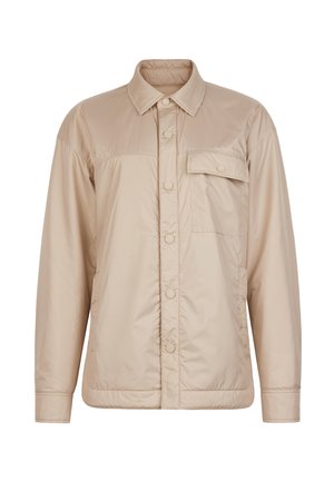 Kobieta o długich brązowych włosach, ubrana w warstwowe beżowe i niebieskie kurtki z zamkiem, stoi na tle jednolitego neutralnego tła, patrząc przed siebie.