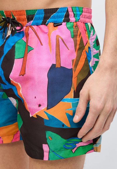 Κοντινό πλάνο ενός άνδρα που φοράει πολύχρωμα μαγιό με αφηρημένα σχέδια, μαύρο κορδόνι και ραφές, με το χέρι να ακουμπάει στο μηρό.