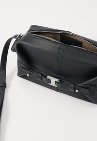 Bolsa de mão preta em couro com fecho em zip, apresentando um detalhe de laço e ferragens prateadas. Textura suave, forma retangular compacta.