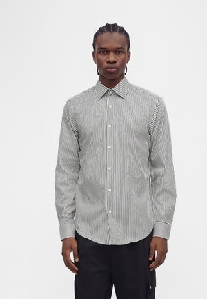 Jeune homme aux cheveux tressés portant une chemise noire et blanche à rayures verticales et manches longues, ainsi qu'un pantalon noir, debout devant un fond uni.