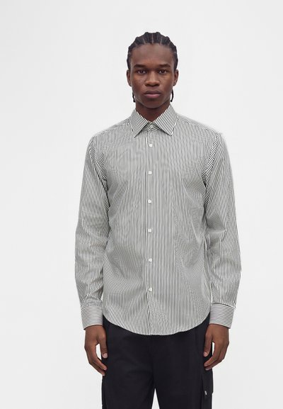 Jeune homme aux cheveux tressés portant une chemise noire et blanche à rayures verticales et manches longues, ainsi qu'un pantalon noir, debout devant un fond uni.