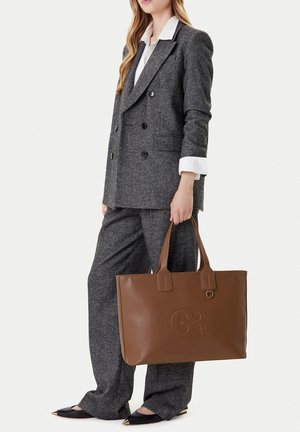 Mujer con traje de pantalón gris y camisa blanca sosteniendo un gran bolso tote de cuero marrón con logo en relieve.
