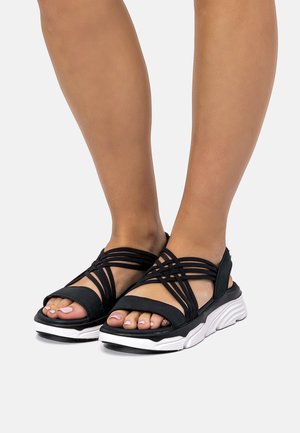 Füße in schwarzen, mit dicken weißen Sohlen versehenen Riemchensandalen, Zehen sichtbar, auf hellem Bodenhintergrund.