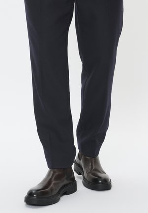 Braune Leder-Chelsea-Stiefel mit glänzender Oberfläche, kombiniert mit dunkelblauen Hosen in maßgeschneiderter Passform, die aus einem strukturierten Stoff gefertigt sind.