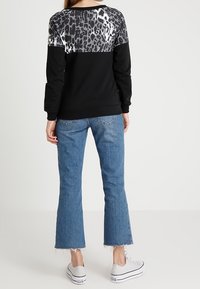 Donna che indossa un felpa nera con yoke a stampa leopardata argentata, jeans blu cropped con orlo sfrangiato e sneakers bianche con plateau, in piedi di spalle.