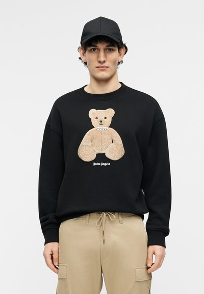 Νέος φοράει μαύρο καπέλο, μαύρο φούτερ με σχέδιο αρκουδάκι σε μπεζ χρώμα και το κείμενο «Palm Angels», καθώς και μπεζ παντελόνι cargo.